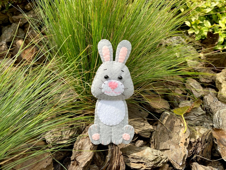 May include: A gray felt bunny with pink ears and nose, sitting on a bed of brown wood chips.