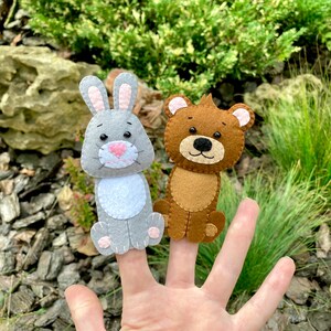 May include: Two felt finger puppets, a gray rabbit and a brown bear, are worn on a person's fingers. The puppets have embroidered eyes and noses.