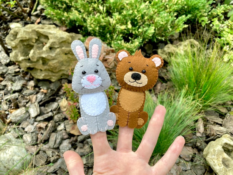 May include: Two felt finger puppets, a gray rabbit and a brown bear, are worn on a person's fingers. The puppets have embroidered eyes and noses.