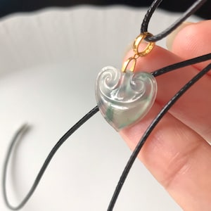 May include: A heart-shaped pendant necklace with a delicate, carved design. The pendant is made of translucent green jade and is set in gold. The necklace is made of black cord.