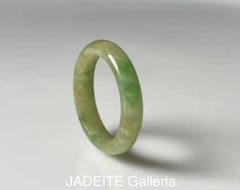 May include: A smooth, polished band ring crafted from natural jadeite stone. The ring features a variegated appearance with swirling patterns of pale cream, light beige, and vibrant emerald green hues. The text JADEITE Galleria is visible.