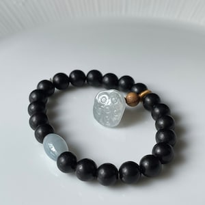 Puede incluir: Un brazalete de cuentas negras con un dije de búho azul claro y una cuenta de madera. El brazalete está sobre una superficie blanca.