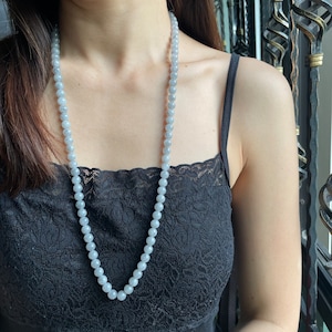 May include: A long necklace made of light gray beads. The necklace is worn by a person with dark hair and is centered in the image.