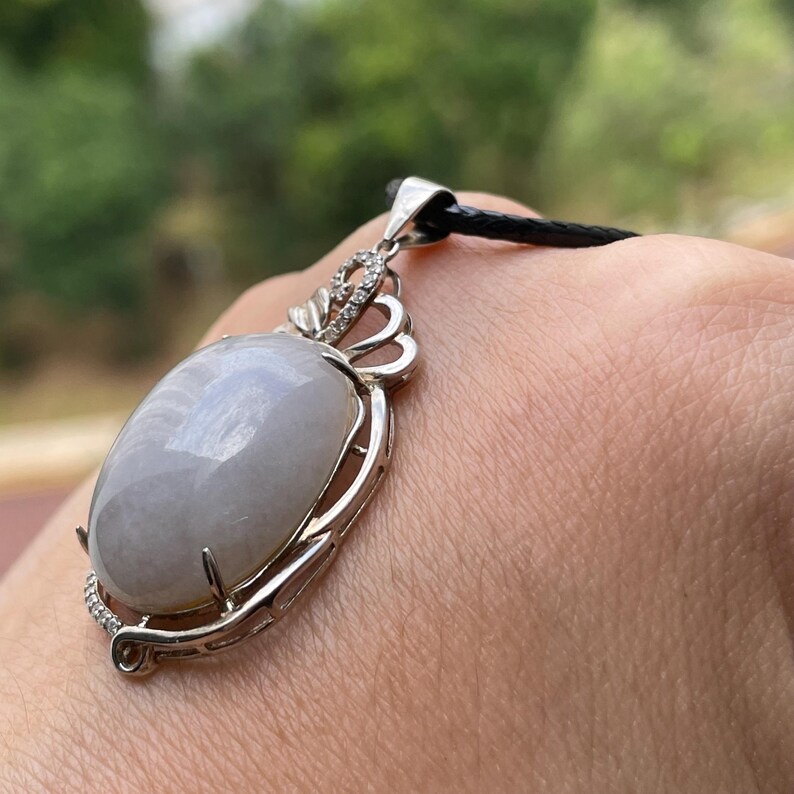 Puede incluir: Un colgante ovalado gris claro con un marco plateado y un cord&oacute;n negro. El colgante tiene una parte superior decorativa plateada con peque&ntilde;as piedras transparentes. El fondo es verde borroso.