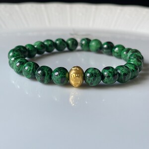 Könnte beinhalten: Ein Armband aus grünem Stein mit einer goldenen Perle in der Mitte. Die Perlen sind klein und rund, und das Armband ist aus Naturstein.