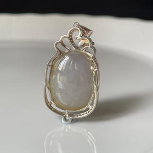 Puede incluir: Colgante de plata con una piedra ovalada transl&uacute;cida, verde p&aacute;lido y blanca. El colgante tiene un marco de plata calado y ornamentado con una parte superior decorativa. La piedra es lisa y pulida, reflejando la luz.