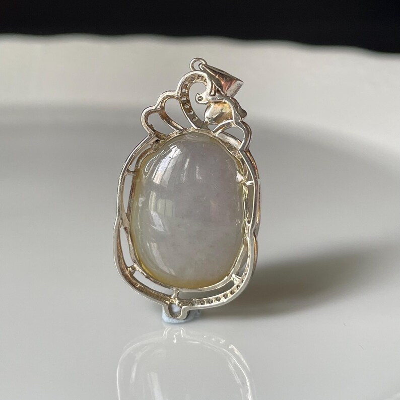 Puede incluir: Colgante de plata con una piedra ovalada transl&uacute;cida, verde p&aacute;lido y blanca. El colgante tiene un marco de plata calado y ornamentado con una parte superior decorativa. La piedra es lisa y pulida, reflejando la luz.