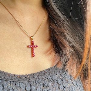 May include: A gold-colored necklace with a cross pendant. The cross is adorned with numerous red gemstones. The necklace is worn against a gray lace top.