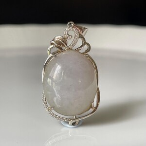 Puede incluir: Colgante ovalado con una piedra preciosa verde p&aacute;lido engastada en un marco de metal plateado. El marco presenta un remolino decorativo en la parte superior y una l&iacute;nea de peque&ntilde;as piedras transparentes a lo largo de la curva inferior. Joyer&iacute;a.