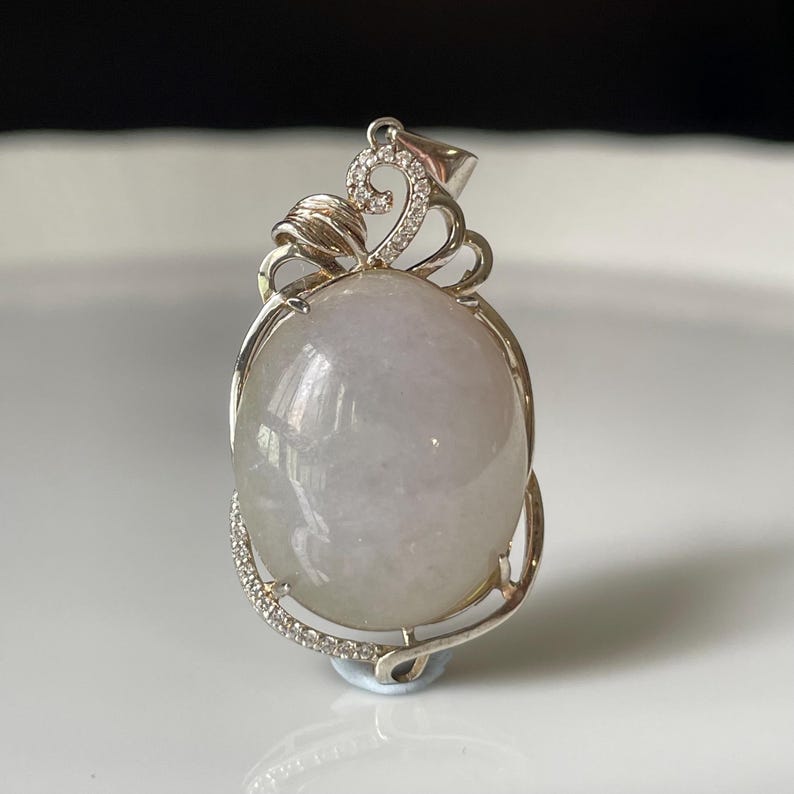 Puede incluir: Colgante ovalado con una piedra preciosa verde p&aacute;lido engastada en un marco de metal plateado. El marco presenta un remolino decorativo en la parte superior y una l&iacute;nea de peque&ntilde;as piedras transparentes a lo largo de la curva inferior. Joyer&iacute;a.