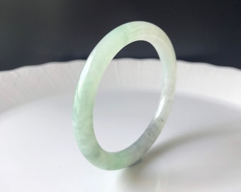 May include: A smooth, polished bangle bracelet crafted from pale green jade stone. The circular accessory features a translucent quality with subtle, natural white and light green mottled patterns throughout its solid, rounded band structure.