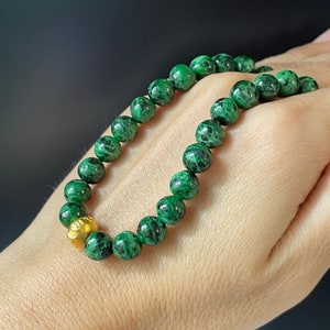 Könnte beinhalten: Ein grünes und schwarzes Edelstein-Armband mit einem goldenen Perlenakzent. Das Armband besteht aus runden Perlen und ist auf einer Schnur aufgefädelt.