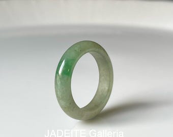 May include: A smooth, polished jadeite ring featuring a translucent pale green body with a concentrated patch of vibrant emerald green color. The band has a rounded profile and a glossy finish. The text JADEITE Galleria is printed below.