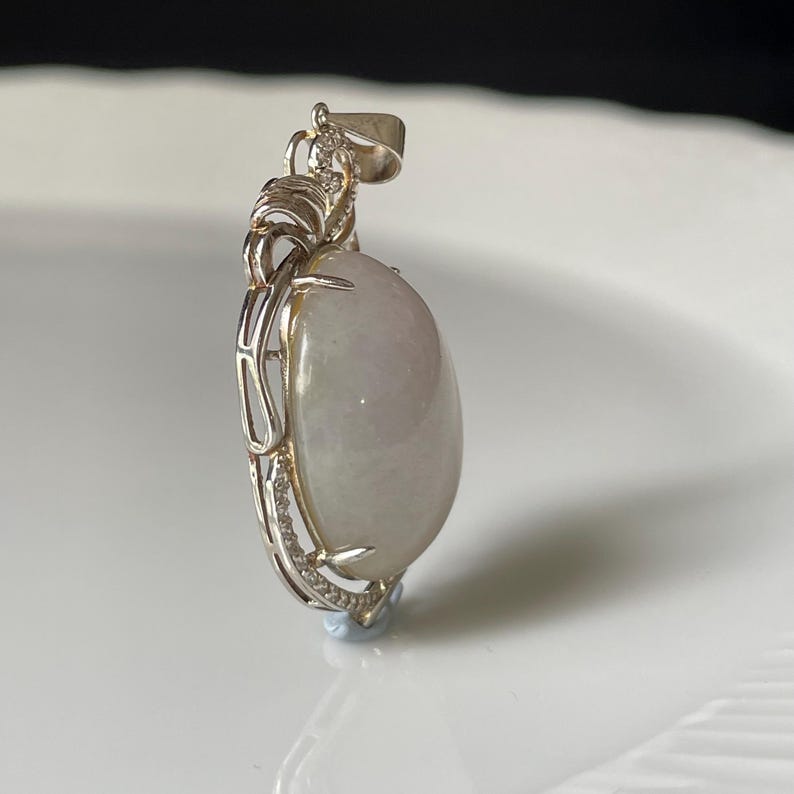 Puede incluir: Colgante de plata con una gran piedra preciosa ovalada de color verde p&aacute;lido. El colgante tiene un marco decorativo de plata con peque&ntilde;as piedras transparentes. La piedra preciosa tiene una superficie lisa y pulida y un sutil degradado de color.