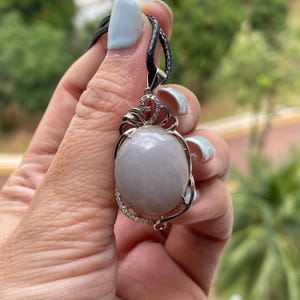 Puede incluir: Collar con colgante ovalado con una piedra preciosa de color lavanda p&aacute;lido. El colgante est&aacute; engastado en un marco de metal plateado, adornado con peque&ntilde;as piedras transparentes y una parte superior decorativa. El collar tiene un cord&oacute;n negro.