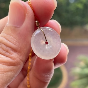 Puede incluir: Un colgante de jade blanco en forma de disco plano con un agujero en el centro, colgado de una cadena de oro.