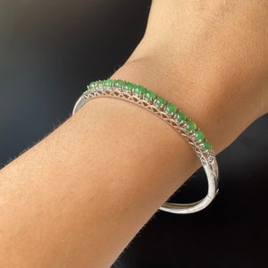 Könnte beinhalten: Ein silbernes Armreif-Armband mit einer Reihe grüner Edelsteine. Das Armband ist offen und hat einen Scharnierverschluss.