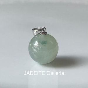 Puede incluir: Un colgante redondo de jade con una argolla de plata. El jade es de color verde claro con un toque de blanco.