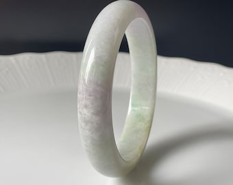 May include: A white jade bangle with a subtle green and purple hue. The bangle is smooth and polished, with a slightly oval shape.