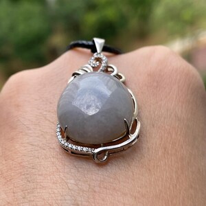 Puede incluir: Un colgante de piedra preciosa gris con un engaste plateado y un cord&oacute;n negro. La piedra preciosa redonda tiene una superficie facetada. El engaste plateado est&aacute; adornado con peque&ntilde;as piedras transparentes. El colgante lo sostiene una persona.