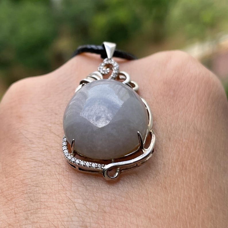 Puede incluir: Un colgante de piedra preciosa gris con un engaste plateado y un cord&oacute;n negro. La piedra preciosa redonda tiene una superficie facetada. El engaste plateado est&aacute; adornado con peque&ntilde;as piedras transparentes. El colgante lo sostiene una persona.