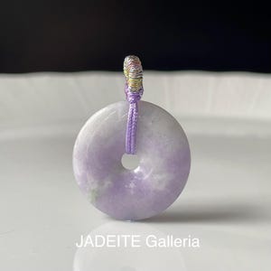 May include: A round, lavender jade pendant with a central hole, suspended by a purple cord. The pendant has a polished finish and a marbled appearance. The text "JADEITE Galleria" is visible at the bottom.