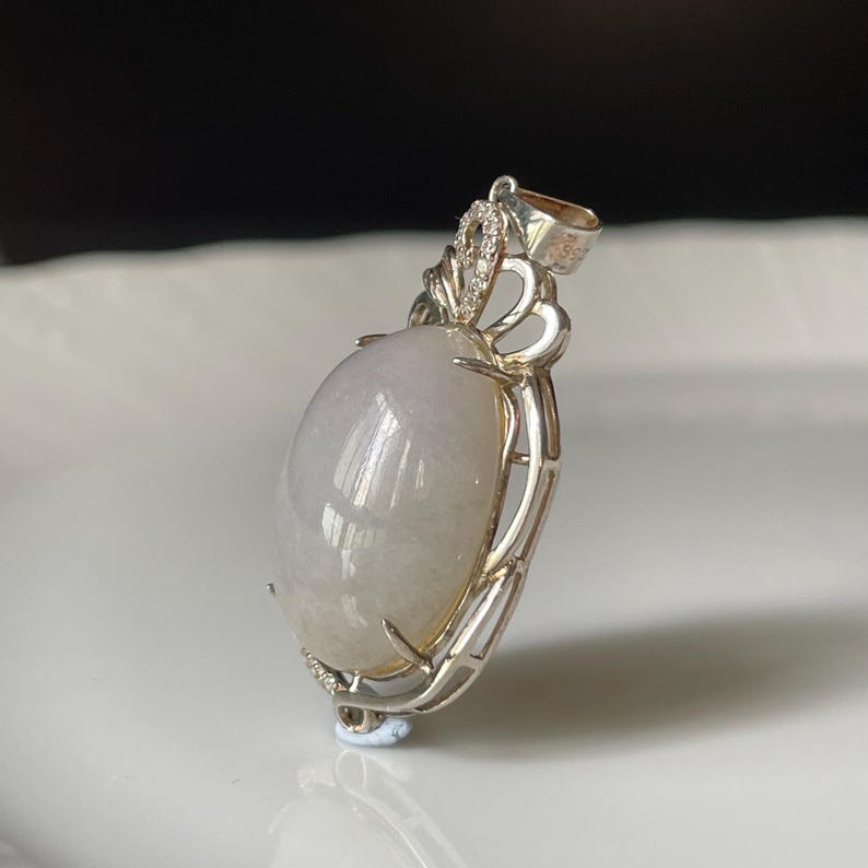 Puede incluir: Colgante de plata con una gran piedra preciosa ovalada de color lavanda p&aacute;lido. La piedra est&aacute; engastada en un marco de plata decorativo con una argolla en la parte superior. La argolla est&aacute; adornada con peque&ntilde;as piedras transparentes.