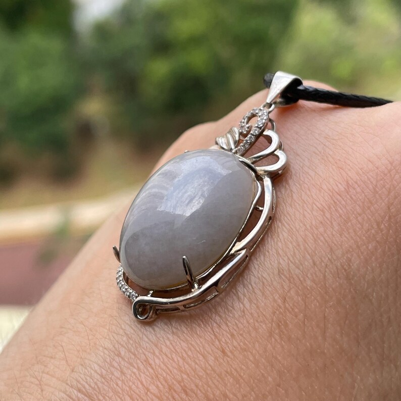 Puede incluir: Un colgante de plata con una piedra grande, ovalada y gris p&aacute;lido. La piedra est&aacute; engastada en un marco de plata decorativo con peque&ntilde;as piedras transparentes. El colgante cuelga de un cord&oacute;n negro.
