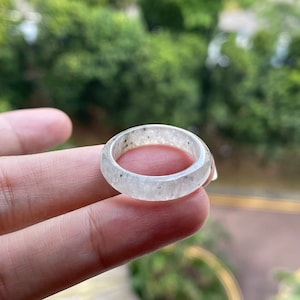 May include: A white jade ring with a speckled pattern. The ring is shown on a finger.