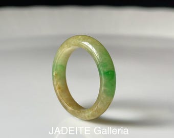 May include: A smooth, circular ring crafted from polished jadeite stone. The band features a gradient of translucent light green and warm amber-toned hues throughout its surface. The text JADEITE Galleria is printed in white at the bottom.