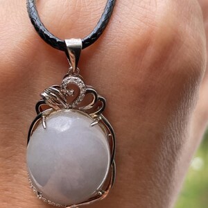 Puede incluir: Un collar con colgante de plata con una gema redonda grande de color azul gris&aacute;ceo p&aacute;lido. El colgante est&aacute; engastado en un marco de plata decorativo con un peque&ntilde;o acento brillante. El collar tiene un cord&oacute;n negro trenzado.