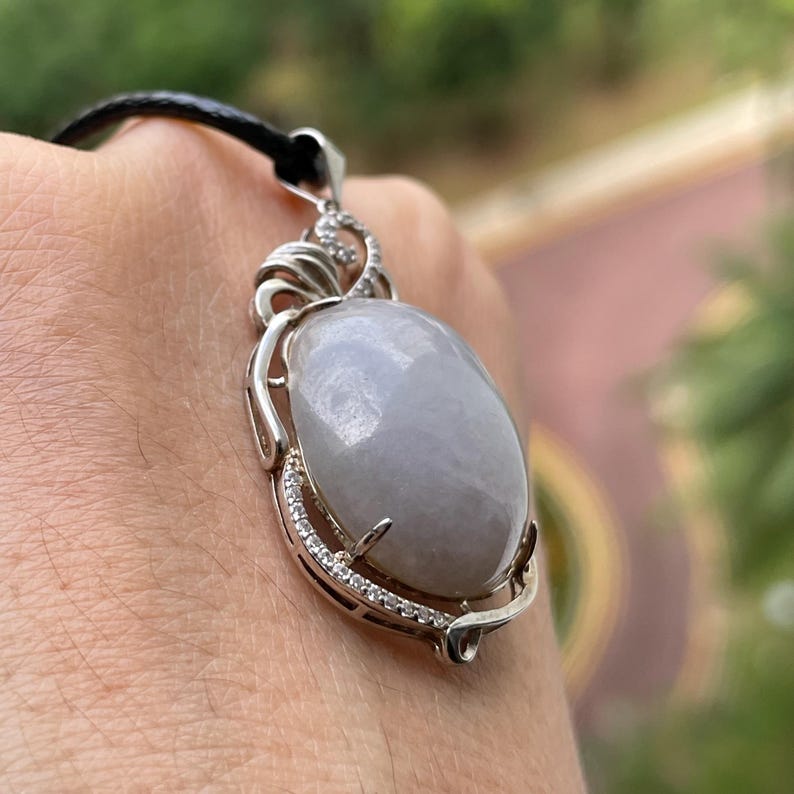 Puede incluir: Un collar con colgante de plata con una gran piedra preciosa ovalada de color gris p&aacute;lido. La piedra preciosa est&aacute; engastada en un marco de plata decorativo con peque&ntilde;as piedras de acento transparentes. El collar tiene un cord&oacute;n negro.