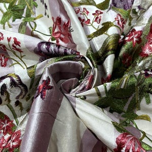 Puede incluir: Primer plano de una tela con un estampado floral. La tela presenta un fondo blanco con rayas verticales lavanda. Flores bordadas en tonos rojos, morados y verdes están dispersas por la tela. La tela tiene una textura brillante, similar al satén.