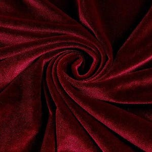 Może przedstawiać: Zbliżenie na głęboki bordowy aksamit. Tkanina jest skręcona i złożona, tworząc spiralny wzór pośrodku. Pluszowy materiał ma miękki połysk, odbijający światło i podkreślający bogaty kolor.