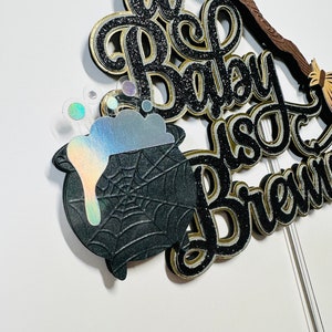 May include: A black and gold glitter cake topper with a cauldron, a broom, and the text "A Baby Is Brewing".
