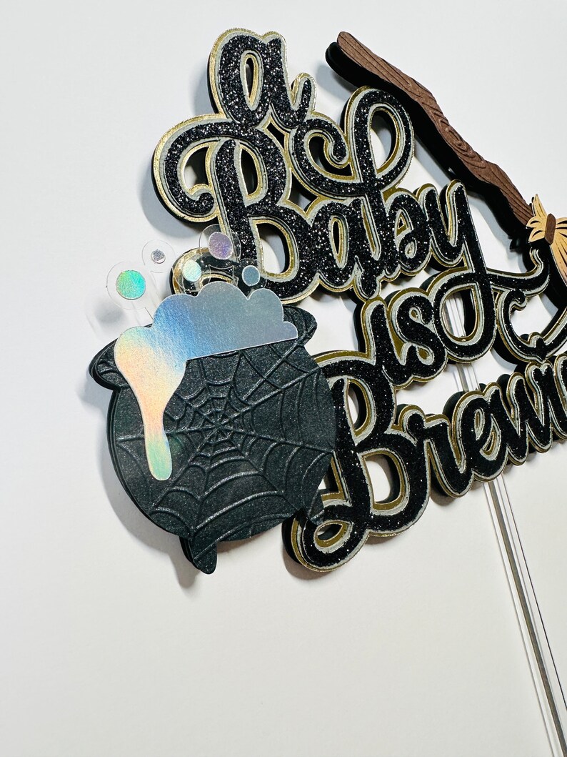 May include: A black and gold glitter cake topper with a cauldron, a broom, and the text "A Baby Is Brewing".