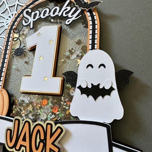 May include: A spooky Halloween cake topper with a white ghost, a black bat, a number one, and the word "Spooky" in black letters. The topper also has a banner with the name "Jack" in orange letters.