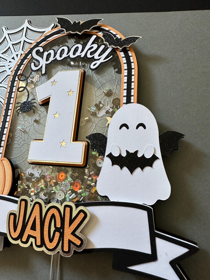 May include: A spooky Halloween cake topper with a white ghost, a black bat, a number one, and the word "Spooky" in black letters. The topper also has a banner with the name "Jack" in orange letters.