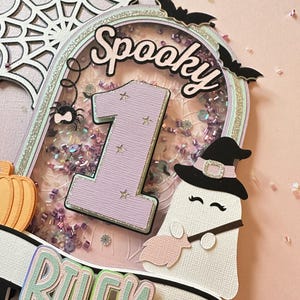 May include: A pink and purple cake topper with a ghost holding a broom, a spider, a pumpkin, a number one, and the words "Spooky" and "Riley".