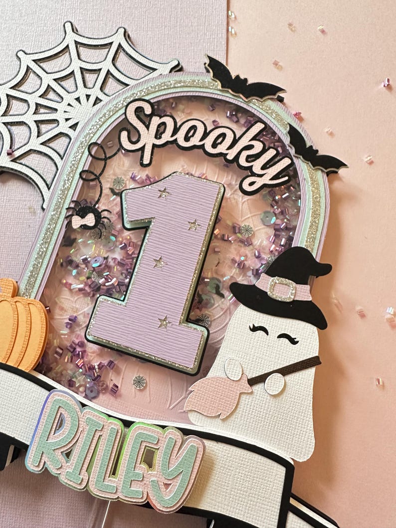 May include: A pink and purple cake topper with a ghost holding a broom, a spider, a pumpkin, a number one, and the words "Spooky" and "Riley".