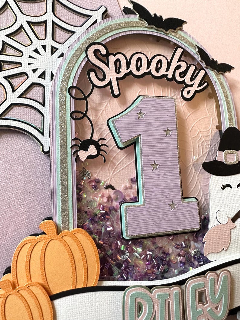 May include: A purple and pink glittery cake topper with a spider web, a spider, a ghost, a pumpkin, and the words "Spooky" and "Riley" on it. The number "1" is in the center of the topper.