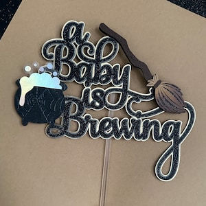 May include: A black and gold glitter cake topper with the text "A Baby is Brewing" and a black cauldron with a spiderweb design and a brown broom.