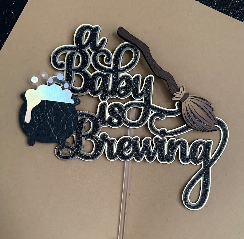 May include: A black and gold glitter cake topper with the text "A Baby is Brewing" and a black cauldron with a spiderweb design and a brown broom.