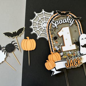 May include: A spooky Halloween cake topper with a white ghost, black spider, and orange pumpkins. The topper features a glittery background with the number "1" and the word "Spooky" in black letters. A white banner with the name "Jack" is attached to the bottom.