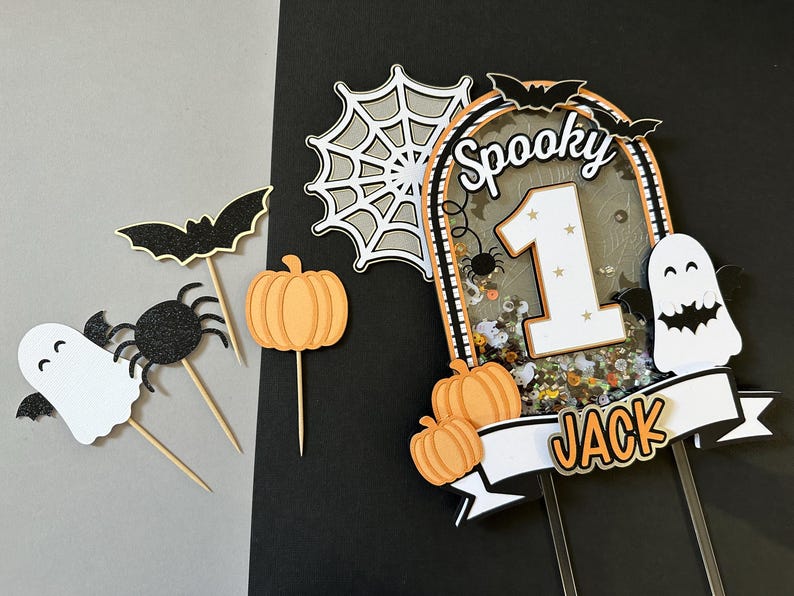 May include: A spooky Halloween cake topper with a white ghost, black spider, and orange pumpkins. The topper features a glittery background with the number "1" and the word "Spooky" in black letters. A white banner with the name "Jack" is attached to the bottom.