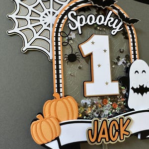 May include: A spooky Halloween cake topper with a white number one, black and white bats, a spider, and a ghost. The topper also features a white banner with the name "JACK" in orange letters and two orange pumpkins.