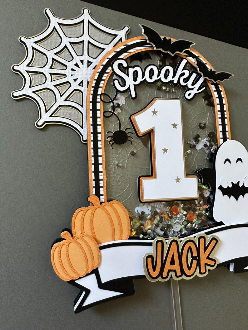 May include: A spooky Halloween cake topper with a white number one, black and white bats, a spider, and a ghost. The topper also features a white banner with the name "JACK" in orange letters and two orange pumpkins.