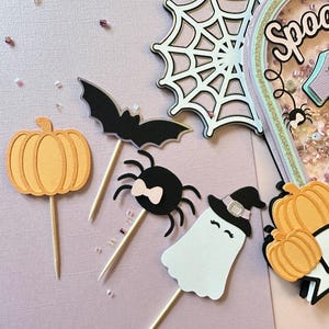 May include: A set of Halloween-themed cake toppers featuring a pumpkin, a spider with a pink bow, a ghost wearing a witch's hat, and a bat. The toppers are made of paper and have wooden sticks for inserting into cakes.