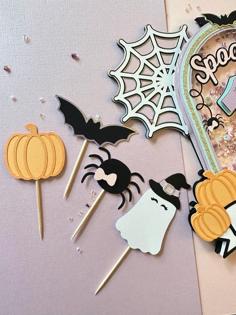 May include: A set of Halloween-themed cake toppers featuring a pumpkin, a spider with a pink bow, a ghost wearing a witch's hat, and a bat. The toppers are made of paper and have wooden sticks for inserting into cakes.