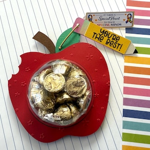 May include: A red apple-shaped gift box filled with gold-wrapped chocolates. The box has a bite taken out of it and is decorated with small stars. A pencil-shaped tag reads "You're the Best!" and a sign says "It Takes a Special Heart to Teach Special Minds."
